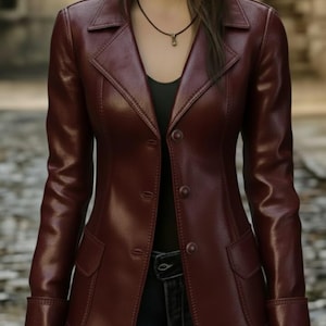 May include: A burgundy leather jacket with a classic design. The jacket features a notched collar, button closure, two front pockets, and long sleeves with buttoned cuffs. The jacket is worn over a black top and blue jeans.