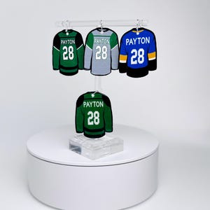 Pode incluir: Miniaturas de camisolas de hóquei em verde, cinza e azul, cada uma com o nome "Payton" e o número "28". As camisolas são exibidas numa plataforma branca e numa haste transparente, mostrando o seu design e detalhes.