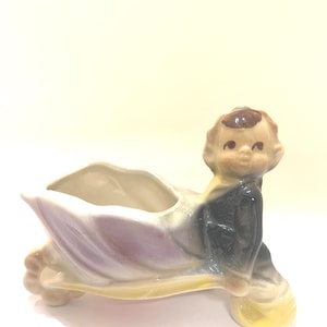 May include: A vintage ceramic planter in the shape of a seated cherub. The cherub has brown hair and is wearing a dark blue robe. The planter is a light cream color with purple and yellow accents.