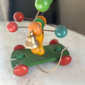 May include: A vintage wooden toy car with a green base and red wheels. The car features a yellow and orange central structure with a small bell and colorful spheres. A wire extends from the car.