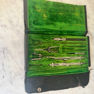 May include: An open, vintage black leather case with a bright green velvet interior. The case contains several silver-colored drafting tools, including compasses and dividers. The case is resting on a white marble surface.