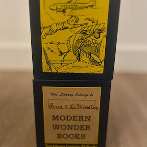 May include: A black and yellow book box with the text "This Library Belongs to Fred e Le Master MODERN WONDER BOOKS This Library Contains 25 Books". The box has a yellow cover with a black and white illustration of a plane, a tiger, and a ship.