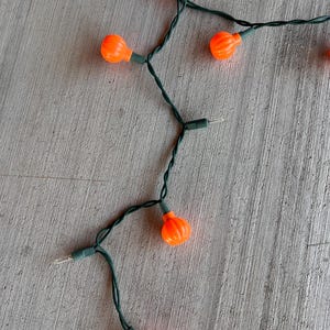 May include: A string of decorative lights featuring small, bright orange pumpkin-shaped covers attached to a dark green electrical cord. The set includes several pumpkin bulbs spaced along the wire, interspersed with clear, unlit bulb sockets.