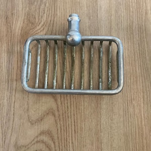 May include: A silver soap dish with a decorative knob on top. The dish has a rectangular shape and a wire grid design.