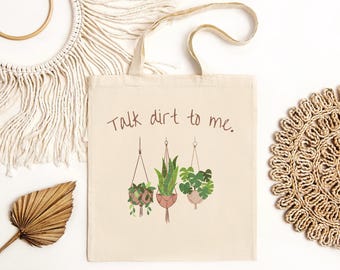 May include: A beige canvas tote bag with the text "Talk dirt to me." printed in brown. The bag features a graphic of three hanging plants with green leaves.
