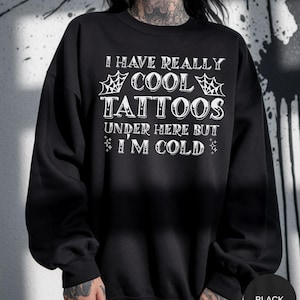 Może przedstawiać: Czarna bluza z białym napisem: "I HAVE REALLY COOL TATTOOS UNDER HERE BUT I'M COLD". Tekst jest ozdobiony pajęczynami i gwiazdami. Bluza jest w jednolitym czarnym kolorze.