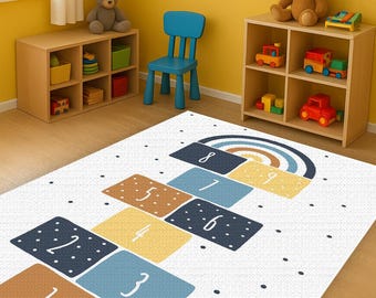 Puede incluir: Una alfombra blanca con un diseño de rayuela con cuadrados numerados en marrón, azul y amarillo. Un arcoíris está encima de los cuadrados. La alfombra está en una sala de juegos para niños con estanterías de madera, juguetes y una silla azul.