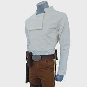 May include: A long-sleeved off-white jacket with a high collar and an asymmetrical front closure, paired with brown trousers featuring a vertical gold-colored stripe down the side. A brown utility belt with a metallic buckle and holster is worn.