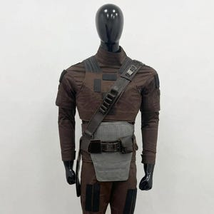 May include: A brown costume on a mannequin. The outfit features a long-sleeved top, trousers, and a utility belt with a grey panel. A shoulder strap with pouches crosses the chest. The costume has black accents and a futuristic design.