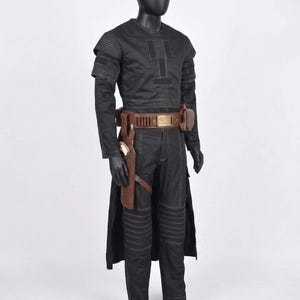 Op de afbeelding: Een zwart kostuum tentoongesteld op een mannequin. De outfit bevat een top met lange mouwen, een broek en een riem met zakjes. Een holster met een pistool is aan het been bevestigd. Het kostuum heeft een futuristisch ontwerp met zichtbare stiksels en gewatteerde details.