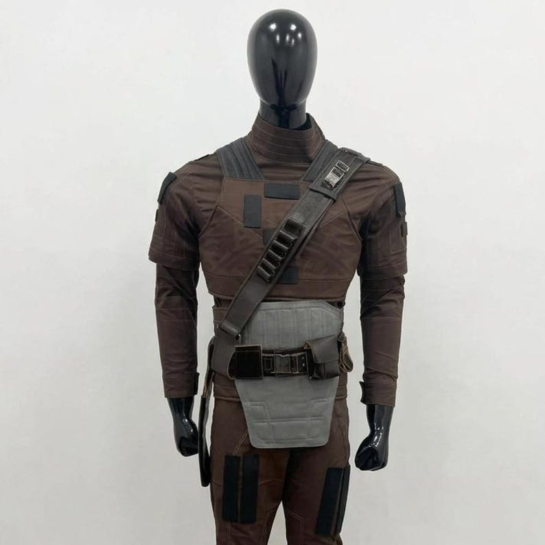 May include: A brown costume on a mannequin. The outfit features a long-sleeved top, trousers, and a utility belt with a grey panel. A shoulder strap with pouches crosses the chest. The costume has black accents and a futuristic design.