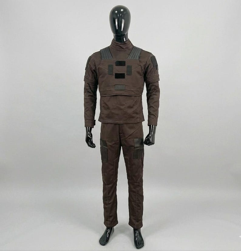 Op de afbeelding: Een bruin tactisch uniform gepresenteerd op een mannequin. De outfit bevat een jas met lange mouwen met versterkte schouder- en borstpanelen, en een bijpassende broek met zakken. De mannequin heeft een zwarte kop en handen.
