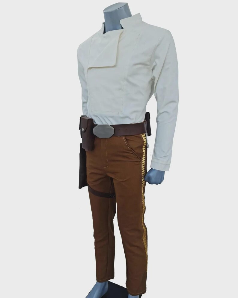 May include: A long-sleeved off-white jacket with a high collar and an asymmetrical front closure, paired with brown trousers featuring a vertical gold-colored stripe down the side. A brown utility belt with a metallic buckle and holster is worn.