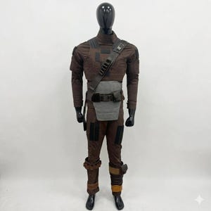 Op de afbeelding: Een bruin kostuum gepresenteerd op een zwarte mannequin. Het kostuum bevat een top met lange mouwen, een broek en diverse accessoires zoals een riem, schouderriem en beenbeschermers. De outfit heeft een tactische uitstraling met meerdere zakken en panelen.
