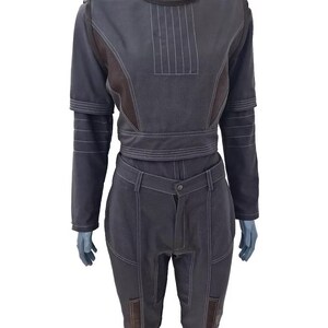 May include: A dark gray two-piece outfit on a mannequin. The top has a high collar, long sleeves, and white stitching details. The pants are high-waisted with matching stitching and brown patches on the knees. The outfit is made of a sturdy material.
