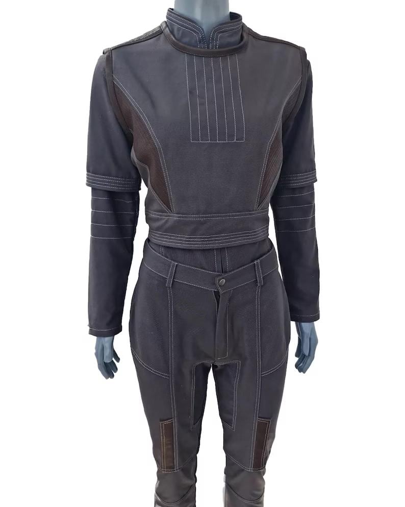 May include: A dark gray two-piece outfit on a mannequin. The top has a high collar, long sleeves, and white stitching details. The pants are high-waisted with matching stitching and brown patches on the knees. The outfit is made of a sturdy material.