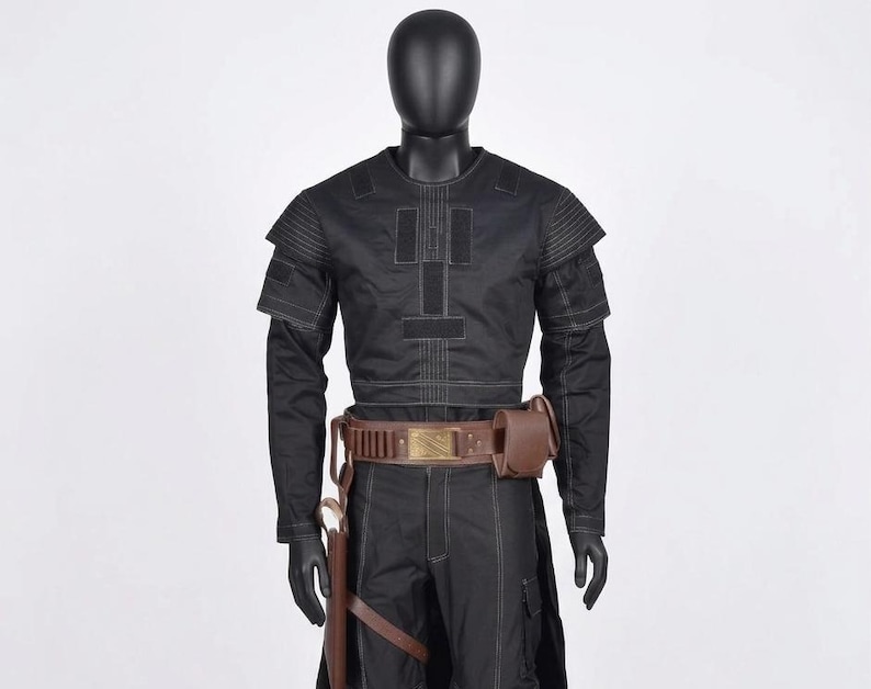 Op de afbeelding: Zwart kostuum met een top met lange mouwen, een broek en een bruine riem met een tasje. De top heeft een geometrisch ontwerp met witte stiksels. Het kostuum wordt op een mannequin gepresenteerd. De outfit bevat een holster en een bruine leren riem.