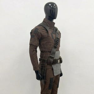 Op de afbeelding: Een bruin tactisch uniform gepresenteerd op een mannequin. De outfit bevat een overhemd met hoge kraag, een broek en een gereedschapsriem met zakken. Een schouderband en een grijs paneel zijn ook zichtbaar. De mannequin heeft een zwarte kop en handen.
