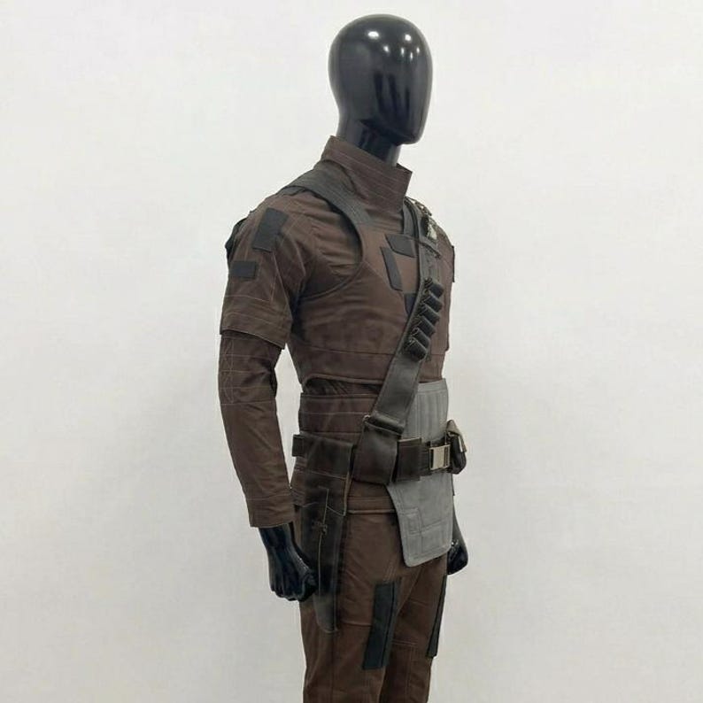 Op de afbeelding: Een bruin tactisch uniform gepresenteerd op een mannequin. De outfit bevat een overhemd met hoge kraag, een broek en een gereedschapsriem met zakken. Een schouderband en een grijs paneel zijn ook zichtbaar. De mannequin heeft een zwarte kop en handen.