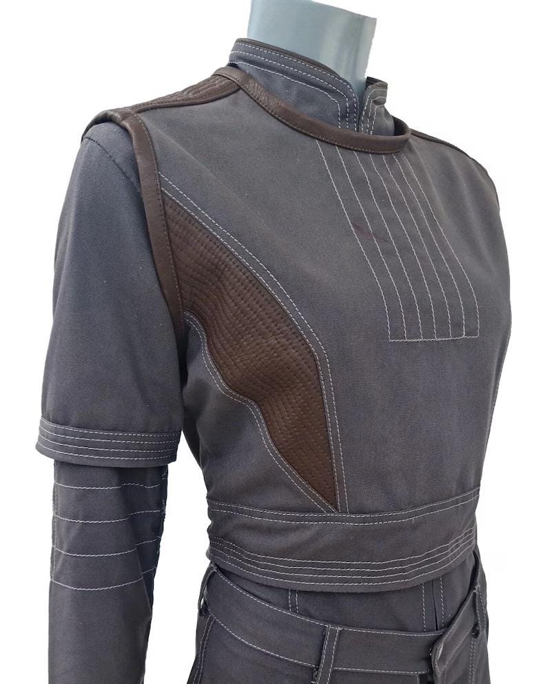 May include: A gray jumpsuit with brown leather accents and white stitching. The jumpsuit features a high collar, short sleeves, and a belted waist. The brown leather is quilted on the shoulders and chest.