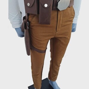 May include: Brown trousers paired with a dark brown leather utility belt featuring two rectangular pouches, a silver-toned hexagonal buckle, and a matching brown leather holster attached to the belt and thigh with a strap.