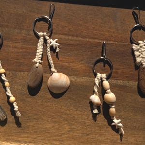 May include: Four handcrafted keychains with natural elements. Each keychain features a silver ring, macrame cord, and unique pendants like driftwood, shells, and wooden beads. The keychains are arranged on a wooden surface, showcasing their rustic charm.