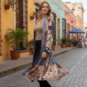 May include: A long, flowing kimono-style robe with a patchwork design in shades of blue, beige, and floral patterns. The robe is worn over a neutral-colored top and black pants. The background features colorful buildings and a cobblestone street.