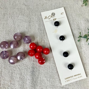 May include: A collection of buttons in various colors and styles. There are several translucent lavender buttons, a cluster of red buttons, and a card of four black buttons. The card reads "Le Chic" and "Guaranteed Washable".