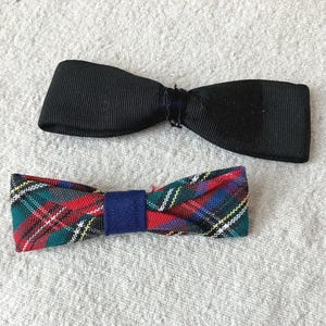 May include: Two bow ties, one solid black with a ribbed texture and a central knot, and one featuring a multi-colored plaid pattern in red, green, blue, and white with a solid blue central band, both constructed from fabric material.