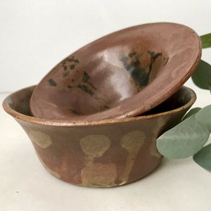 May include: Two stacked ceramic bowls with a smooth, reddish-brown glazed finish. The bottom bowl features a series of light tan, rounded vertical shapes around its exterior, while the top bowl has dark, mottled black markings on its interior.