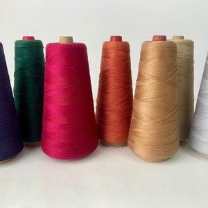 May include: A collection of eight spools of thread in various colors, including deep purple, emerald green, and vibrant red. The spools are arranged in a row, showcasing the range of hues available for sewing and crafting projects.