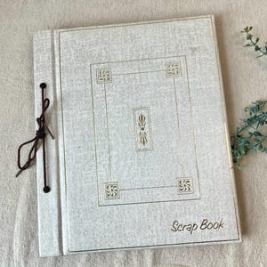 May include: A vintage scrapbook with a light gray cover featuring a gold geometric design and the words "Scrap Book." The book is bound with a dark brown cord and has a decorative design.