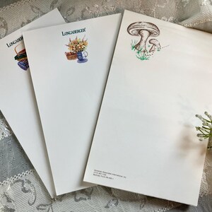 May include: Three white Longaberger notepads with different illustrations. One notepad features a mushroom design, another has a basket and flowers, and the third has a basket and a blue cup with flowers. The Longaberger logo is at the top of each notepad.