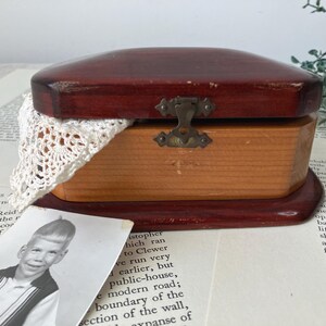 May include: A vintage wooden jewelry box with a hinged lid, stained in a deep reddish-brown. The box has a light-colored wooden base and a brass clasp. A white lace doily and a black and white photo are visible.