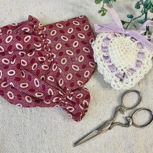 May include: A burgundy bonnet with a white floral pattern, a white crocheted heart with lavender ribbon, and ornate silver scissors are arranged on a white surface. The heart is decorated with lavender ribbon.