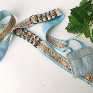 May include: A light blue ribbon belt featuring a floral pattern with pink and green accents. The belt includes two sections of bronze-toned metal rings woven with the floral ribbon and a small, light blue square pouch attached to one end.