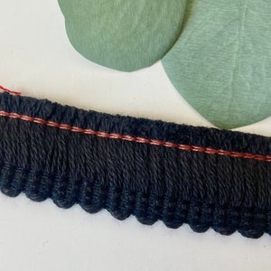 May include: A strip of black fringe trim featuring a dense, textured weave with a single horizontal line of metallic copper-colored stitching running across the upper portion. The bottom edge of the trim is finished with a series of small, rounded loops.