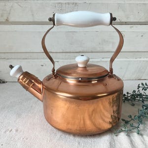 May include: A polished copper tea kettle featuring a rounded body, a curved spout with a white ceramic tip, and a matching white ceramic handle. The lid is topped with a small white ceramic knob, and the metal frame connects the handle to the base.