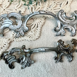 May include: Two ornate, antique-style metal drawer pulls with a distressed silver finish. The pulls feature intricate scrollwork and a curved handle design, perfect for adding vintage charm to furniture.