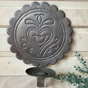 May include: A gray metal wall sconce with a scalloped edge and a heart design. The heart and decorative elements are created with punched holes. The sconce has a small, round candle holder attached below.