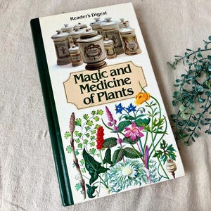 May include: A hardcover book titled "Magic and Medicine of Plants" by Reader's Digest. The cover features illustrations of various plants and apothecary jars. The book has a white cover with a green spine.