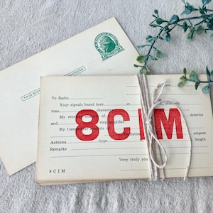 May include: Vintage radio log cards tied with twine, featuring the bold red letters "8 CIM." The cards have handwritten text and are accompanied by a postcard with a green stamp. The background is a light-colored fabric.
