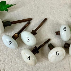 May include: Seven antique white porcelain door knobs with aged metal stems are arranged on a neutral surface. The knobs are numbered 1 through 7. The oval-shaped knobs have a classic, vintage aesthetic, suitable for restoration projects or adding a touch of history.