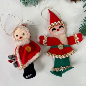 May include: Two vintage felt Christmas ornaments. One is a Santa figure in a red cape with white trim and a black base labeled Japan. The other is a smiling elf figure in a red hat, red tunic with gold trim, and green pants, both with gold hanging loops.