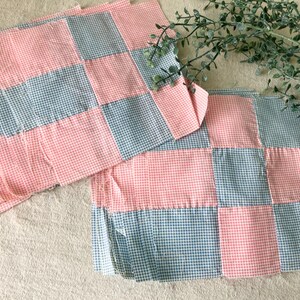 May include: Two patchwork fabric pieces with a gingham pattern in pink and blue. The fabric is arranged in a grid pattern, with alternating squares of pink and blue. The pieces are placed on a neutral-colored surface.
