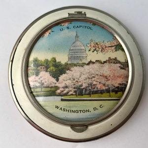 May include: Vintage round metal picture frame featuring a colorful image of the U.S. Capitol building in Washington, D.C., surrounded by cherry blossoms. The frame has a silver finish and the text "U.S. CAPITOL" and "WASHINGTON, D.C." are visible.