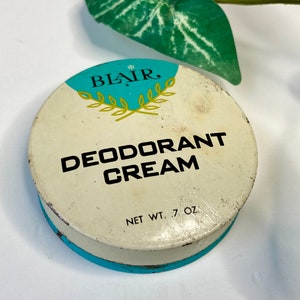 May include: A round, vintage metal tin of Blair Deodorant Cream with a white lid featuring teal accents, a gold laurel leaf graphic, and black text reading BLAIR, DEODORANT CREAM, and NET WT. .7 OZ. The container has visible signs of wear and age.
