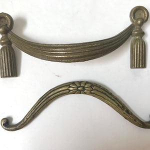 May include: Two antique brass drawer pulls. One is a curved handle with a floral center. The other is a decorative handle with a draped design and tassel accents. Both are made of brass.