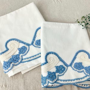 May include: Two folded white fabric cloths featuring a decorative border of blue and cream-colored crochet work. The crochet pattern consists of scalloped edges, floral motifs, and swirling shapes that create a layered, textured trim along the bottom.