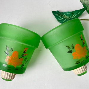 May include: Two green glass flower pots with orange rose and leaf designs. The pots have a frosted finish and a white and beige base. The pots are tilted towards each other.